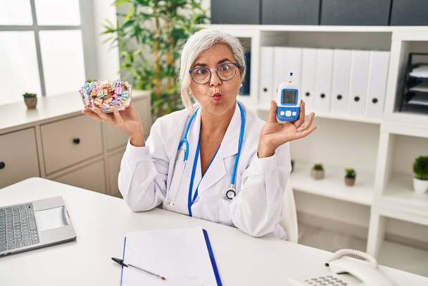 Kobieta w średnim wieku z siwymi włosami, nosząca mundur lekarza, trzymająca monitor glukozy, robiąca rybią twarz ustami i mrugającymi oczami, szalona i komiczna..  - Zdjęcie, obraz