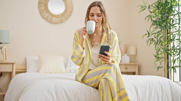 Young blonde woman using smartphone drinking coffee at bedroom - Zdjęcie, obraz