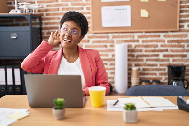 Jovem afro-americana trabalhando no escritório usando óculos sorrindo com a mão sobre o ouvido ouvindo rumores ou fofocas. conceito de surdez.  - Foto, Imagem