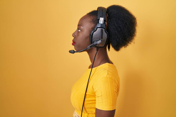 Afrikanerin mit lockigem Haar steht über gelbem Hintergrund und trägt Kopfhörer, die zur Seite schauen, entspannte Profilpose mit natürlichem Gesicht mit selbstbewusstem Lächeln.  - Foto, Bild