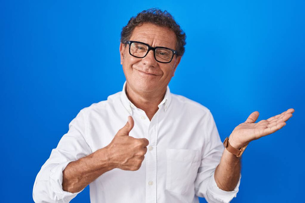 Keski-ikä latino mies seisoo sininen tausta osoittaa kämmen ja tekee ok ele peukalot pystyssä, hymyillen onnellinen ja iloinen  - Valokuva, kuva