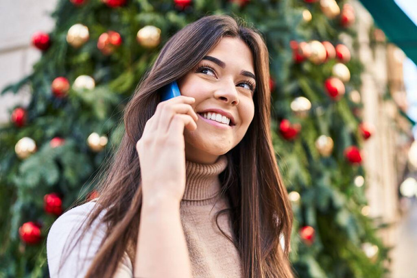 Junge schöne hispanische Frau spricht mit Smartphone, das am Weihnachtsbaum auf der Straße steht - Foto, Bild