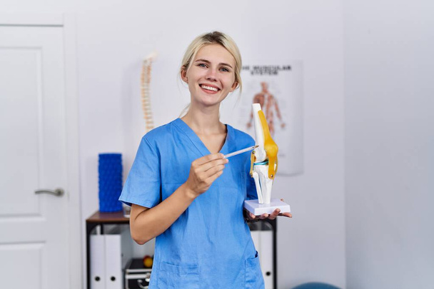 Junge blonde Pysiotherapeutin lächelt selbstbewusst und zeigt auf anatomisches Knie-Modell in Reha-Klinik - Foto, Bild