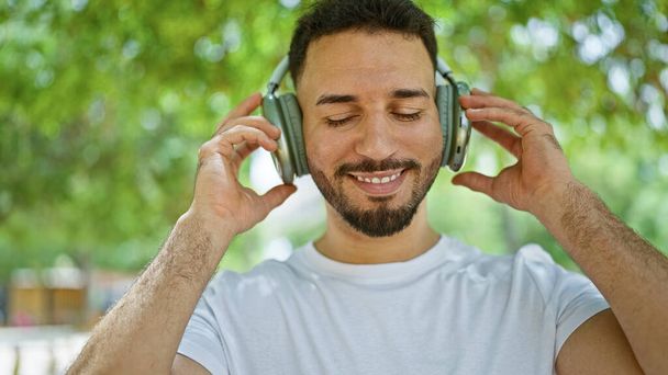 Mladý arabský muž poslouchající hudbu usmívající se v parku - Fotografie, Obrázek