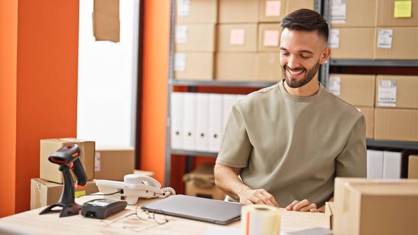 Молодий іспаномовний чоловік електронної комерції бізнес сидить на столі посміхаючись в офісі - Фото, зображення