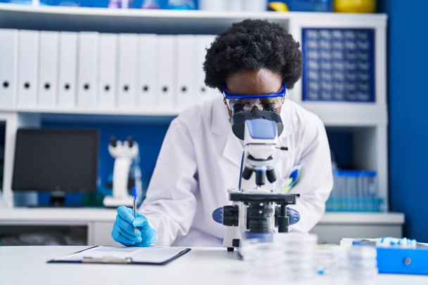 Jonge Afrikaans-Amerikaanse vrouwelijke wetenschapper schrijft op document met behulp van microscoop in het laboratorium - Foto, afbeelding