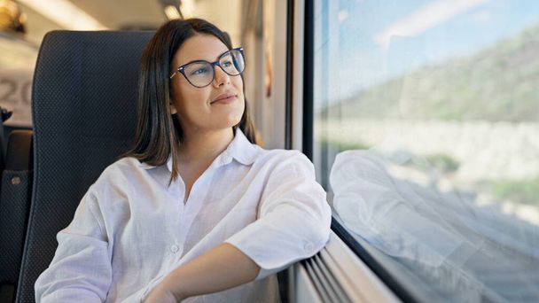 Joven hermosa mujer hispana sonriendo feliz mirando a través de la ventana dentro de vagón de tren - Foto, imagen