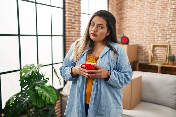 Young beautiful plus size woman drinking coffee with relaxed expression at new home - Foto, imagen