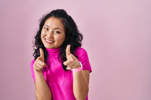 Jeune femme asiatique debout sur fond rose pointant les doigts vers la caméra avec un visage heureux et drôle. bonne énergie et vibes.  - Photo, image