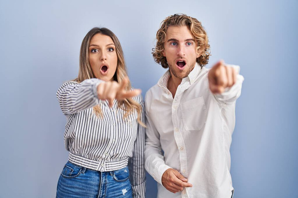 Молодая пара стоит на синем фоне указывая пальцем удивлен вперед, открытый рот удивлен выражение, что-то на передней  - Фото, изображение