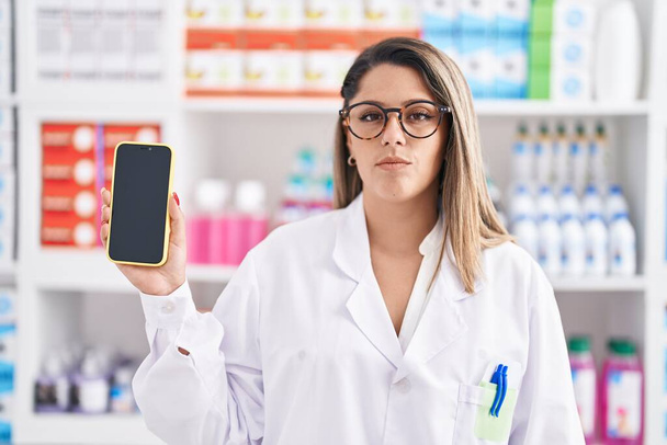 Blondýny žena pracující v lékárně drogerie ukazující smartphone obrazovce myšlení postoj a střízlivý výraz hledá sebevědomí  - Fotografie, Obrázek