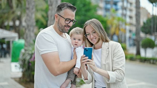 Familie aus Mutter, Vater und Baby lächelt mit Smartphone auf der Straße - Foto, Bild