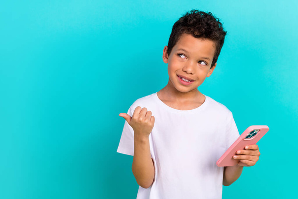 Portrait of small cute schoolboy hold smart phone look indicate thumb finger empty space ad isolated on turquoise color background. - 写真・画像