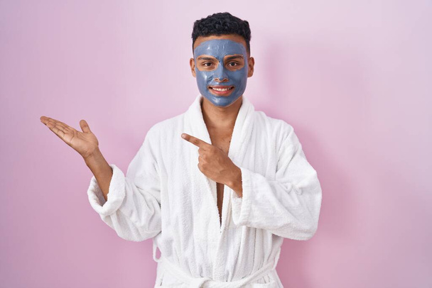 Jonge Spaanse man draagt schoonheid gezichtsmasker en badjas verbaasd en glimlachend naar de camera terwijl presenteren met de hand en wijzen met de vinger.  - Foto, afbeelding