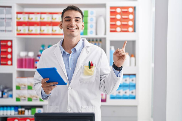 Hezký hispánský muž pracující v lékárně s tabletem šťastně ukazující rukou a prstem na stranu  - Fotografie, Obrázek