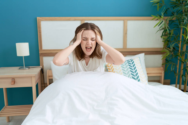 Νεαρή λευκή γυναίκα στρεσαρισμένη να κάθεται στο κρεβάτι ουρλιάζοντας στην κρεβατοκάμαρα - Φωτογραφία, εικόνα