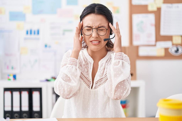Hispanische junge Frau, die im Büro arbeitet, mit Headset und Brille, die Hand auf dem Kopf, Kopfschmerzen wegen Stress. Migräne.  - Foto, Bild