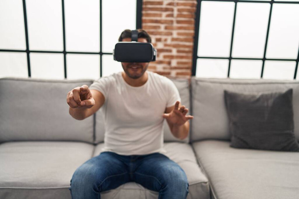 Молодий іспаномовний чоловік грає у відеоігри, використовуючи окуляри віртуальної реальності вдома
 - Фото, зображення