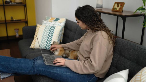 Mladá hispánská žena se psem pomocí notebooku sedí na pohovce doma - Fotografie, Obrázek