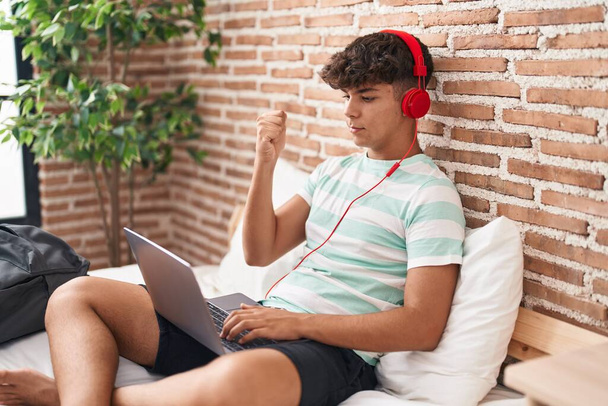 Hispanic teenager using laptop at home sitting on the bedroom screaming proud, celebrating victory and success very excited with raised arms  - Photo, Image