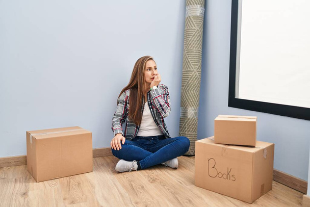 Fiatal szőke nő ül a padlón, költözik egy új otthonba, stresszesnek és idegesnek tűnik, miközben a kezei körmöket harapdálnak. szorongás.  - Fotó, kép