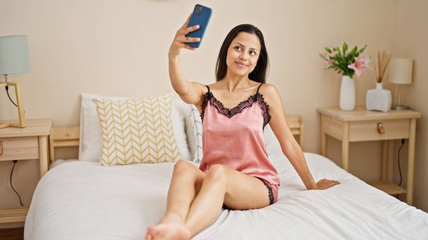 Joven hermosa mujer hispana hacer selfie por teléfono inteligente sentado en la cama en el dormitorio - Foto, imagen