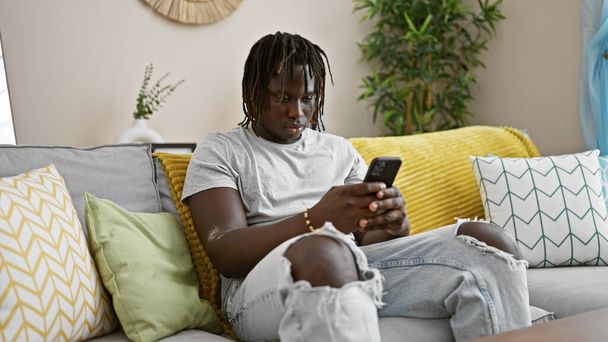 Afrikkalainen amerikkalainen mies käyttää älypuhelinta istuu sohvalla kotona - Valokuva, kuva