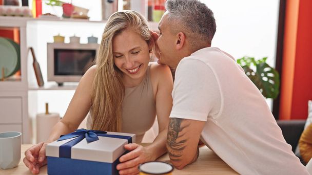 Man and woman couple sitting on table with gift kissing at dinning room - Photo, Image