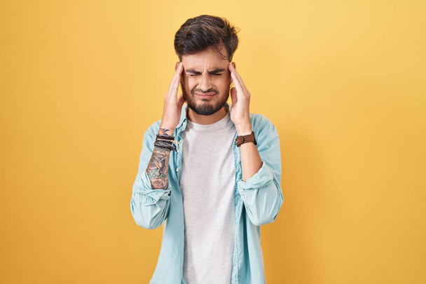 Jeune homme hispanique avec des tatouages debout sur fond jaune avec la main sur la tête, maux de tête parce que le stress. migraine souffrant.  - Photo, image