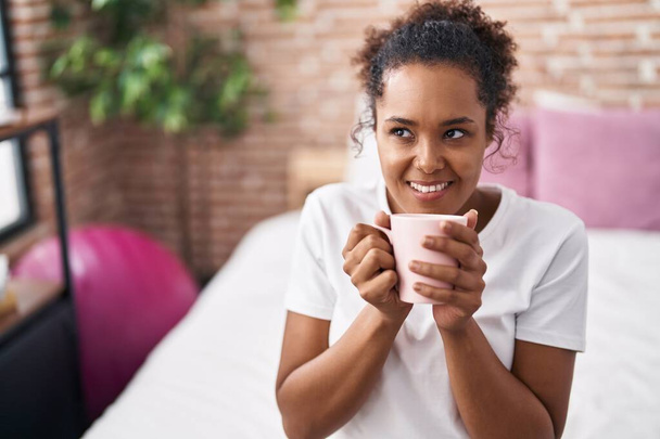 Femme afro-américaine buvant une tasse de café assise sur le lit dans la chambre - Photo, image