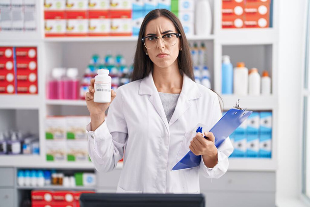 Jeune femme brune travaillant à la pharmacie drugstore tenant des pilules sceptiques et nerveuses, fronçant les sourcils contrariés en raison de problèmes. personne négative.  - Photo, image