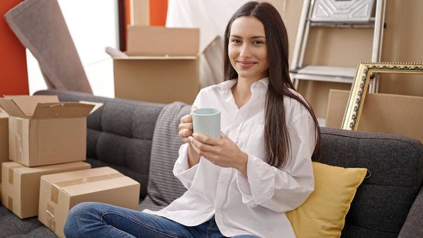 Young beautiful hispanic woman drinking coffee sitting on sofa at new home - Photo, Image