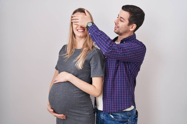 Ein junges Paar erwartet ein Baby, das vor weißem Hintergrund steht, lächelnd und lachend mit der Hand auf dem Gesicht die Augen zur Überraschung verdeckt. Blindes Konzept.  - Foto, Bild
