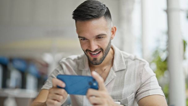 Fiatal spanyol férfi mosolygós magabiztos videojáték a kávézó teraszán - Fotó, kép