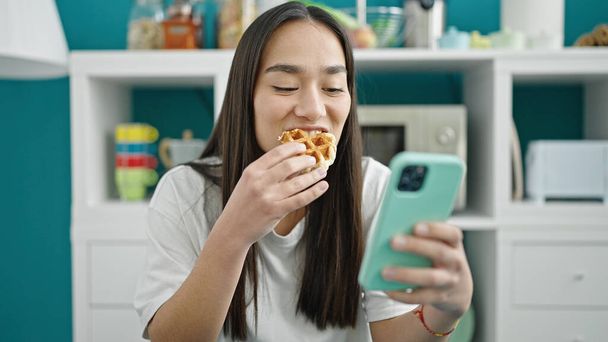Genç ve güzel İspanyol kadın waffle yiyor. Yemek odasında akıllı telefon kullanıyor. - Fotoğraf, Görsel