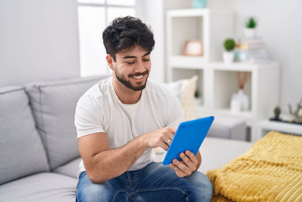Homem hispânico com barba usando touchpad sentado no sofá com um sorriso feliz e legal no rosto. pessoa de sorte.  - Foto, Imagem