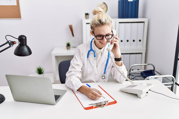 Молодая блондинка врач говорит по телефону письменном виде на документе в клинике - Фото, изображение