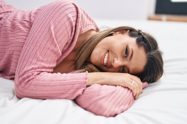 Junge hispanische Frau lächelt selbstbewusst auf Bett im Schlafzimmer liegend - Foto, Bild