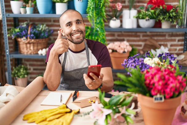 Hombre hispano con barba trabajando en floristería con teléfono inteligente sonriendo feliz y positivo, pulgar hacia arriba haciendo excelente y signo de aprobación  - Foto, imagen