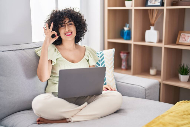 Νεαρή μελαχρινή γυναίκα με σγουρά μαλλιά χρησιμοποιώντας φορητό υπολογιστή κάθεται στον καναπέ στο σπίτι χαμογελώντας θετικά κάνει ok σημάδι με το χέρι και τα δάχτυλα. επιτυχής έκφραση.  - Φωτογραφία, εικόνα