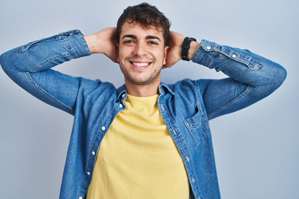 Joven hombre hispano de pie sobre fondo azul relajante y estiramiento, brazos y manos detrás de la cabeza y el cuello sonriendo feliz  - Foto, Imagen