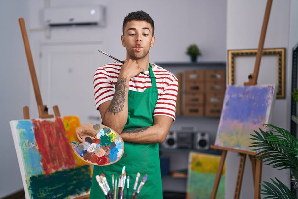 Βραζιλιάνος νεαρός άνδρας κρατώντας παλέτα ζωγράφος στο στούντιο καλλιτέχνη κάνοντας το πρόσωπο ψαριών με το στόμα και τα μάτια squinting, τρελό και κωμικό.  - Φωτογραφία, εικόνα