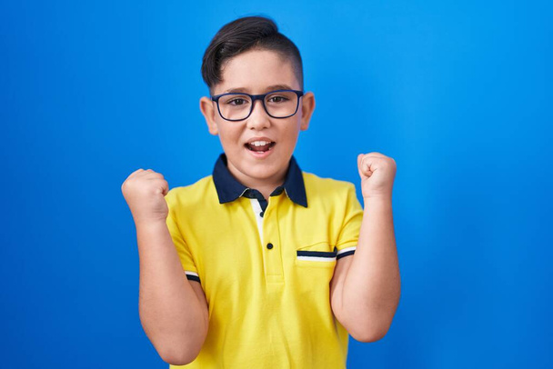 Young hispanic kid standing over blue background screaming proud, celebrating victory and success very excited with raised arms  - Foto, immagini