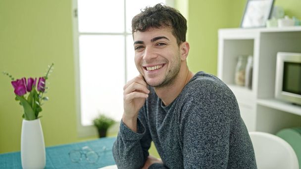 Молодой латиноамериканец, уверенно улыбающийся, сидя на столе в столовой - Фото, изображение
