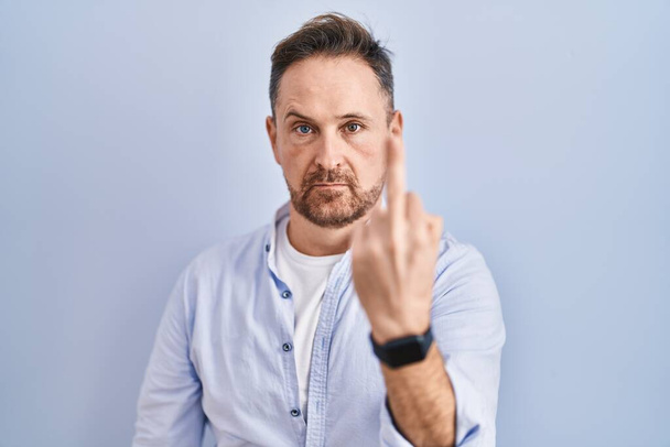 Middle age caucasian man standing over blue background showing middle finger, impolite and rude fuck off expression  - Фото, изображение
