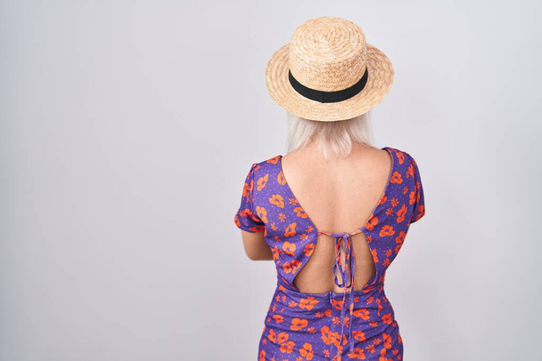 Νεαρή λευκή γυναίκα με λουλουδάτο φόρεμα και καλοκαιρινό καπέλο να στέκεται πίσω κοιτώντας μακριά με σταυρωμένα τα χέρια  - Φωτογραφία, εικόνα