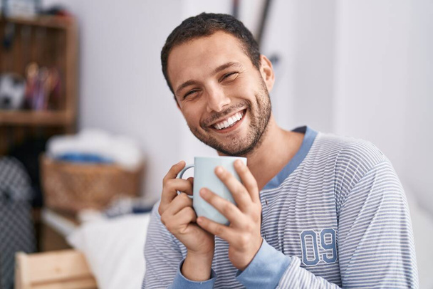 Young man drinking cup of coffee sitting on bed at bedroom - Photo, Image