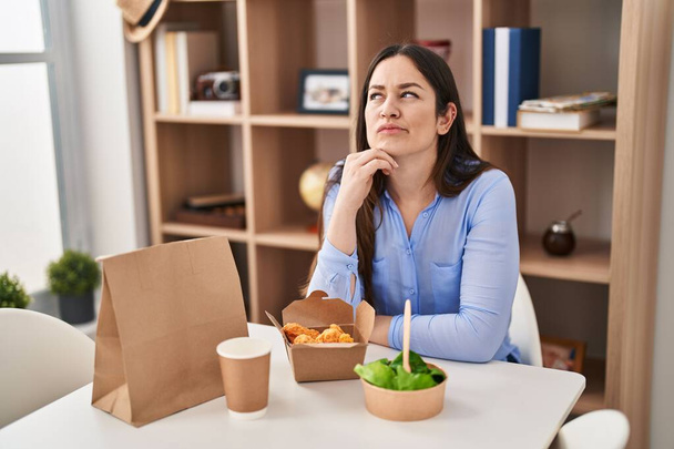 jonge brunette vrouw eten afhaalmaaltijden thuis serieus gezicht denken over vraag met de hand op kin, bedachtzaam over verwarrende idee  - Foto, afbeelding
