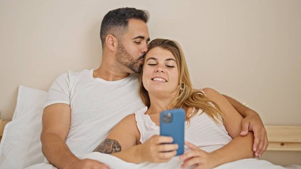 Man and woman couple lying on bed using smartphone hugging each other at bedroom - Photo, Image
