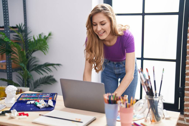Joven mujer rubia artista sonriendo confiado usando ordenador portátil en el estudio de arte - Foto, Imagen